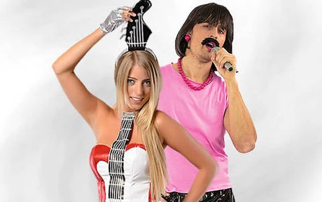 Eine Frau in einem Kleid mit Klaviertasten und einem Gitarren-Stirnband posiert neben einem Mann in einem rosa Hemd und einer schwarzen Perücke, der ein Mikrofon in der Hand hält.