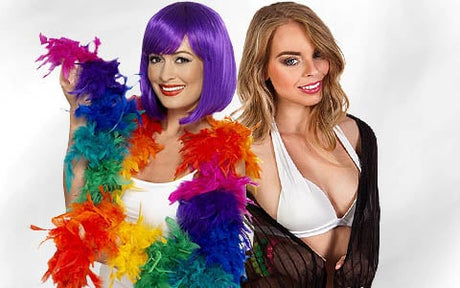 Zwei Frauen posieren gemeinsam; die eine trägt eine lila Perücke und eine Regenbogen-Federboa, die andere einen weißen BH und ein schwarzes Überkleid. Beide lächeln vor einem schlichten weißen Hintergrund.