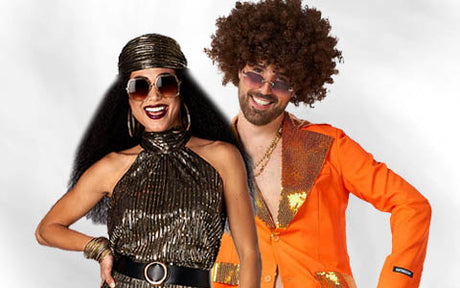 Zwei Personen in Outfits im Stil der 1970er Jahre; die Frau trägt ein Stirnband und ein glänzendes Kleid, und der Mann trägt einen orangefarbenen Anzug und eine Afro-Perücke. Beide lächeln und tragen eine Sonnenbrille.