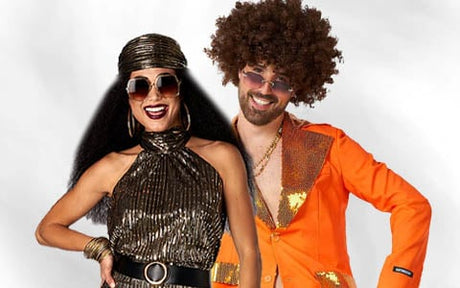 Zwei Personen in Retro-Outfits posieren gemeinsam; die eine trägt ein glitzerndes schwarzes Kleid und ein Stirnband, die andere einen orangefarbenen Anzug mit Pailletten und eine braune Afro-Perücke.