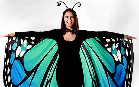 Eine Person in einem Schmetterlingskostüm mit ausgebreiteten schwarzen, blauen und grünen Flügeln, die vor einem weißen Hintergrund steht.