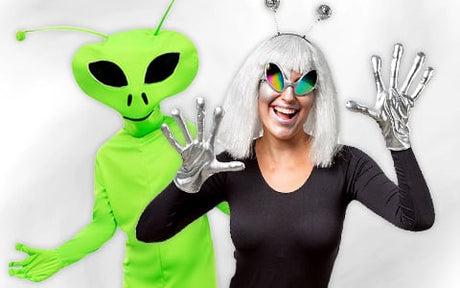 Zwei Personen in Außerirdischenkostümen posieren; eine trägt einen leuchtend grünen Anzug und eine Maske, die andere eine weiße Perücke, ein Stirnband mit Antennen, silberne Handschuhe und eine verspiegelte Brille.