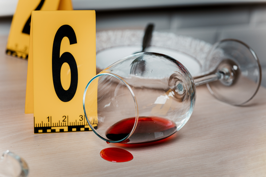 Ein umgestürztes Weinglas mit verschütteter roter Flüssigkeit neben einer gelben Markierung mit der Nummer 6 auf einem Tisch.