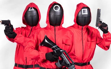 Drei Personen in roten Overalls mit Kapuze und schwarzen Masken, auf denen Dreieck-, Kreis- und Quadratsymbole zu sehen sind, stehen Seite an Seite vor einem schlichten Hintergrund und halten jeweils eine Waffe in der Hand.