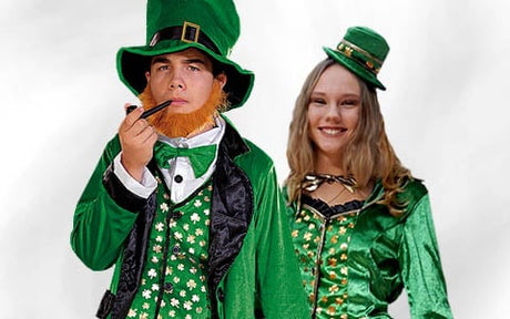 Zwei Personen in grünen Leprechaun-Kostümen, von denen eine eine Pfeife in der Hand hält und einen orangefarbenen Kunstbart trägt; beide stehen vor einem schlichten Hintergrund und blicken in die Kamera.