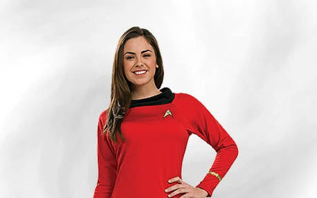 Eine Frau in einem roten „Star Trek“-Uniformkleid steht vor einem schlichten weißen Hintergrund, lächelt und stützt eine Hand in die Hüfte.