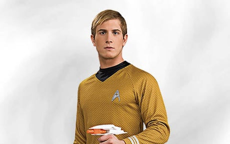 Eine Person, die eine gelbe „Star Trek“-Uniform trägt und einen Phaser in der Hand hält, steht vor einem schlichten, hellen Hintergrund.