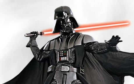 Eine als Darth Vader verkleidete Person hält in einer Hand ein rotes Lichtschwert und streckt den anderen Arm nach vorne aus; sie trägt eine schwarze Rüstung und einen Umhang.