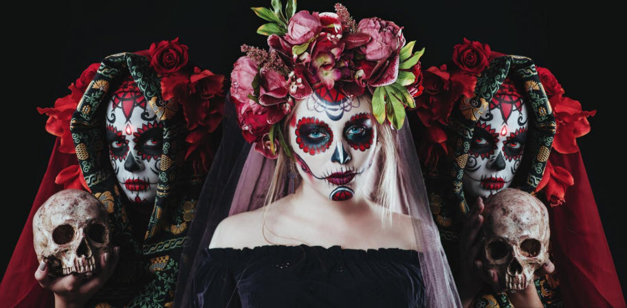 Drei Personen mit bemalten Totenkopfgesichtern und Blumenkränzen posieren vor einem schwarzen Hintergrund; zwei halten Totenköpfe in den Händen, und die Person in der Mitte trägt einen rosa Schleier und einen großen Blumenkranz.