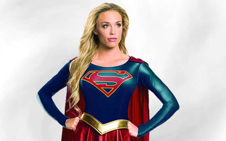 Eine als Supergirl verkleidete Person steht mit den Händen in den Hüften vor einem hellgrauen Hintergrund und trägt ein blau-rotes Kostüm mit einem S-Emblem, einen roten Umhang und einen goldenen Gürtel.