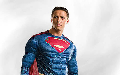 Ein Mann in einem blauen Superman-Kostüm mit rotem Umhang steht vor einem schlichten, hellen Hintergrund und blickt leicht zur Seite.