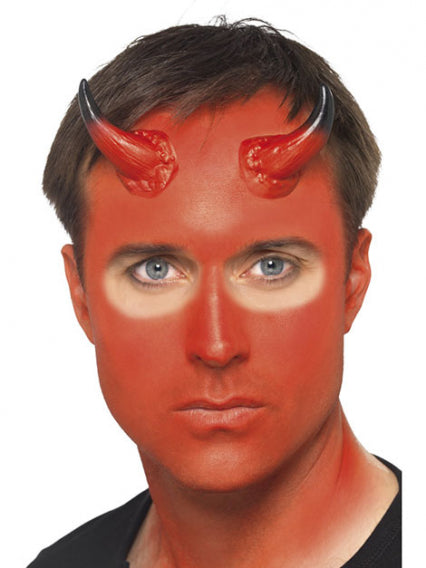 Ein Mann mit roter Gesichtsbemalung, schwarzen Haaren und zwei roten Teufelshörnern auf der Stirn, der direkt in die Kamera blickt.