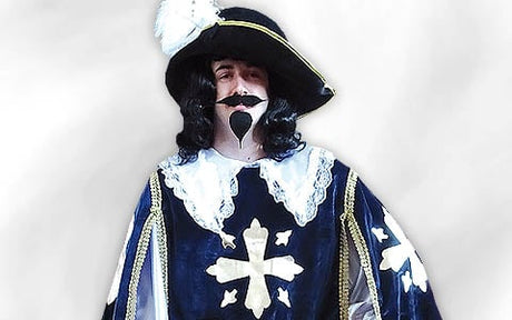 Eine Person in historischer Tracht mit einem Federhut, einer schwarzen Perücke und einem Schnurrbart, die einen marineblauen Umhang trägt, der mit weißen Kreuzen und Spitzenbesatz verziert ist, und vor einem hellen Hintergrund steht.