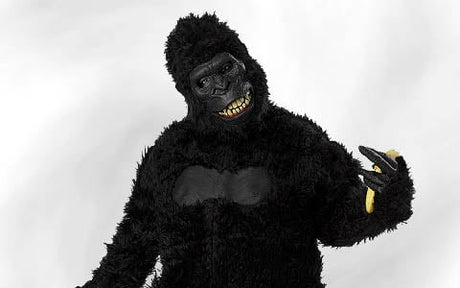 Eine Person in einem schwarzen Gorillakostüm posiert vor einem schlichten weißen Hintergrund und hält in der rechten Hand eine halb geschälte Banane, auf die sie zeigt.