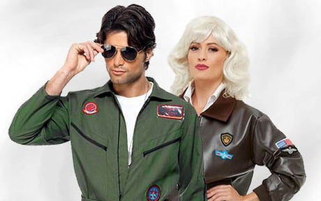 Zwei Personen tragen Kostüme im Pilotenstil; der Mann hat dunkles Haar, trägt eine Sonnenbrille und einen grünen Fluganzug, während die Frau blondes Haar hat, eine braune Jacke trägt und die Hände in die Hüften gestemmt hat.