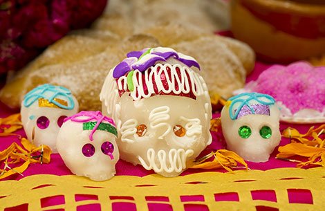 Auf einem farbenfrohen Tisch mit traditionellem Brot sind vier verzierte Zuckerschädel und Ringelblumenblüten ausgestellt, die die Feierlichkeiten zum Día de los Muertos symbolisieren.