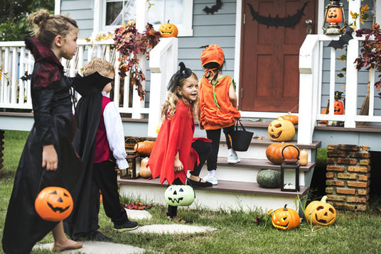 Vier Kinder in Halloween-Kostümen mit Kürbiskörbchen nähern sich einer geschmückten Veranda mit geschnitzten Kürbissen und Herbstblättern.