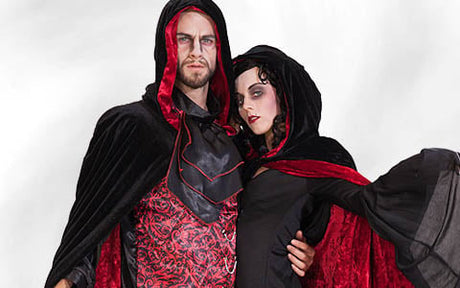 Zwei Personen in schwarzen und roten Kapuzenumhängen posieren gemeinsam vor einem schlichten weißen Hintergrund; sie tragen auffälliges Make-up und Kostüme im Gothic-Stil.