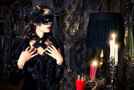 Eine Frau mit einer schwarzen Maskenballmaske und einem schwarzen Kleid steht in einem dunklen, reich verzierten Raum, der mit Spinnweben geschmückt ist, neben brennenden Kerzen, einem Getränk und einer Lampe.