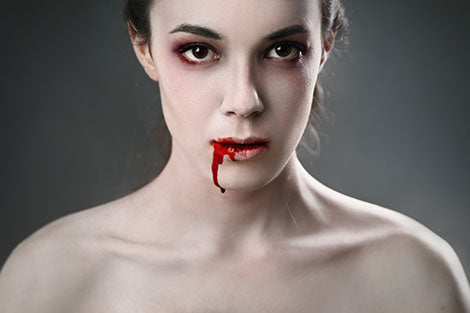 Eine blasse Person mit dunklem Lidschatten, aus deren Mund rotes Blut tropft, blickt vor einem grauen Hintergrund direkt in die Kamera.