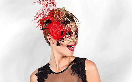 Eine Frau in einem schwarzen Kleid, die eine verzierte rot-goldene Maskenballmaske mit Federn und einer Blume trägt, lächelt und blickt vor einem schlichten Hintergrund zur Seite.