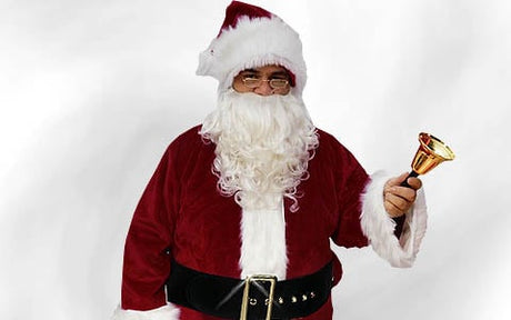 Eine als Weihnachtsmann verkleidete Person, die eine kleine goldene Glocke in der Hand hält, einen roten Anzug, einen weißen Bart, eine Brille und eine rote Mütze trägt und vor einem schlichten weißen Hintergrund steht.