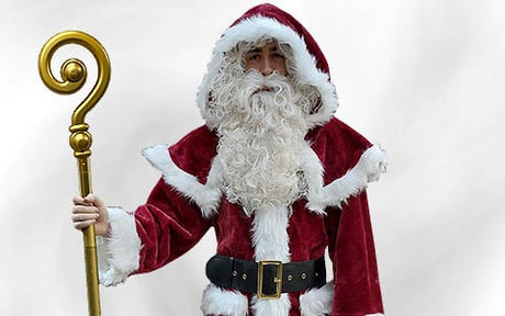Eine Person in einem rot-weißen Kostüm im Weihnachtsmann-Stil mit einem lockigen weißen Bart, die einen goldenen Stab in der Hand hält und vor einem schlichten Hintergrund steht.