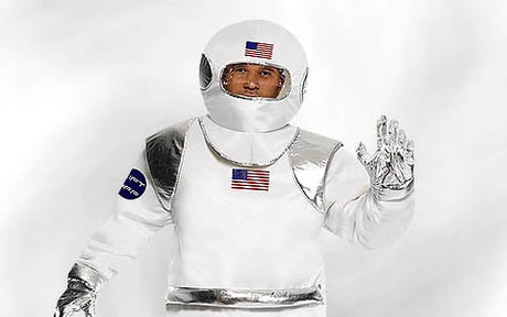 Eine Person in einem weiß-silbernen Raumanzug mit Aufnähern in Form der amerikanischen Flagge, die eine Hand hebt und vor einem schlichten weißen Hintergrund steht.