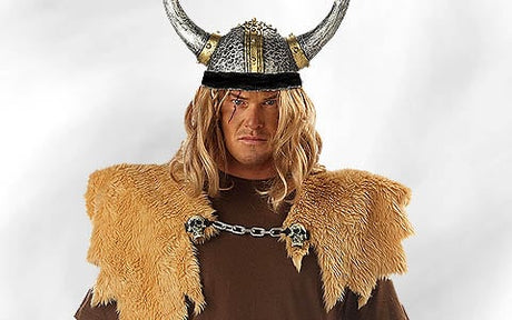 Eine Person mit einem Wikingerhelm mit Hörnern, einer langen blonden Perücke, einem braunen Hemd und einer Schulterbedeckung aus Fell, die vor einem schlichten hellen Hintergrund steht.