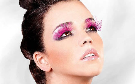 Eine Frau mit glänzenden Lippen und auffälligen rosa, gefiederten Wimpern blickt vor einem weißen Hintergrund nach oben.