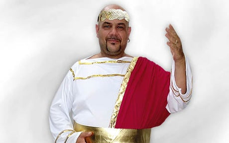 Vor einem schlichten Hintergrund steht ein Mann in einer weißen Toga mit goldenen Verzierungen und einer roten Schärpe, einen Arm erhoben; er trägt ein goldenes Stirnband.