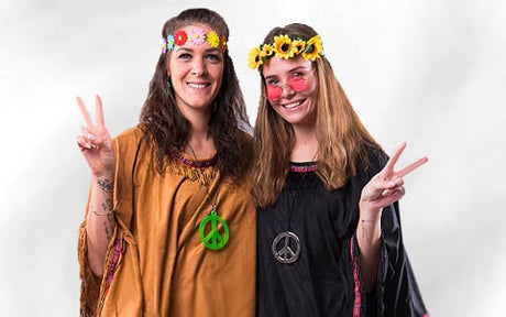 Zwei Frauen in Hippie-Kostümen aus den 1960er Jahren, mit Blumenstirnbändern und Halsketten mit Peace-Zeichen, die beide die Peace-Geste machen und lächeln.