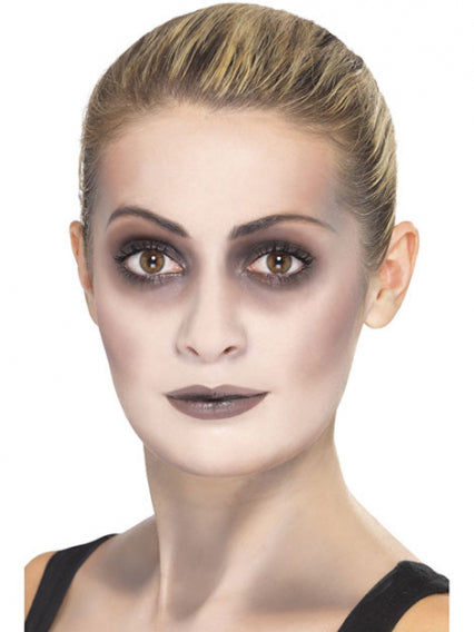 Eine Frau mit dezentem Make-up, dunklen Augenringen und neutraler Lippenfarbe, die vor einem schlichten weißen Hintergrund nach vorne blickt.