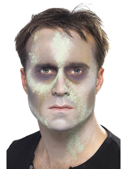 Ein Mann mit kurzen braunen Haaren hat sein Gesicht blass geschminkt, mit grünen und grauen Flecken, was ihm vor dem weißen Hintergrund ein gespenstisches oder zombieähnliches Aussehen verleiht.