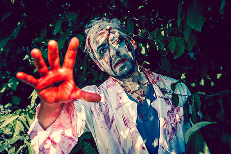 Eine Person in Zombie-Make-up und -Kostüm, die mit einer blutverschmierten Hand nach vorne greift und draußen inmitten von grünem Laub steht.