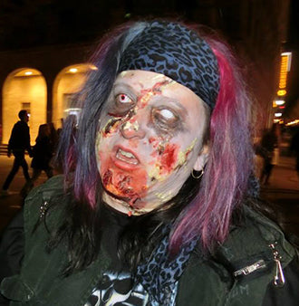 Eine Person mit Zombie-Make-up, künstlichen Wunden, verfärbter Haut und zerzaustem lila Haar, die ein Stirnband und dunkle Kleidung trägt und nachts draußen steht.