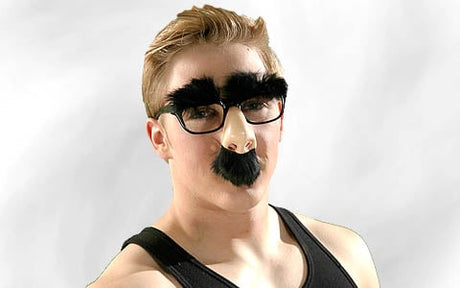 Eine Person, die eine lustige Verkleidung mit dicken schwarzen Augenbrauen, einer falschen Nase, einem Schnurrbart und einer Brille trägt und ein schwarzes Tanktop anhat, vor einem schlichten hellen Hintergrund.
