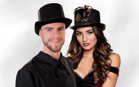 Ein Mann und eine Frau in schwarzen Outfits und Hüten im Steampunk-Stil stehen gemeinsam vor einem schlichten weißen Hintergrund.